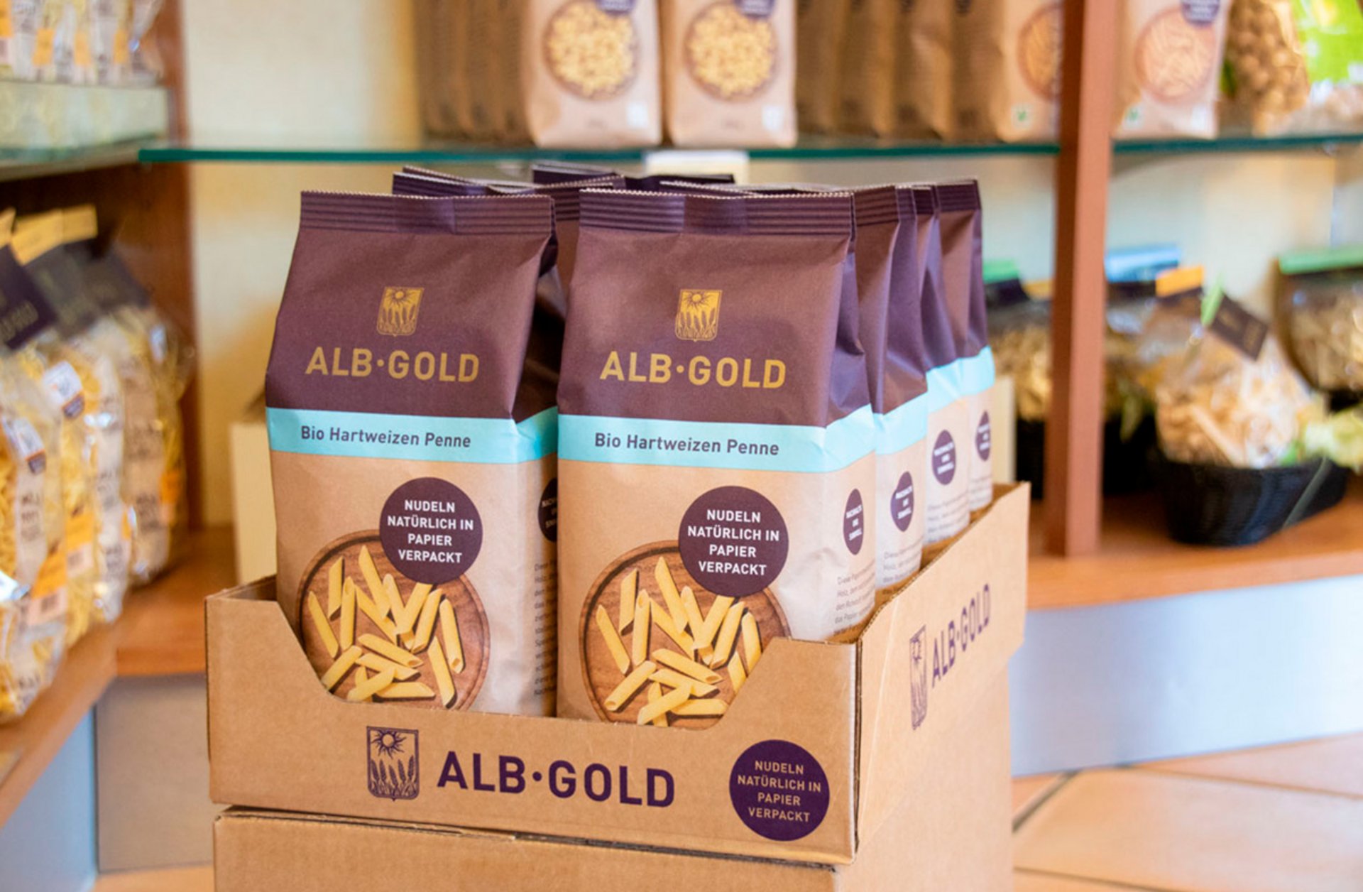 Mehrere Packungen ALB-GOLD Bio Hartweizen Penne aus Papierverpackung stehen in einem Karton auf einem Ladenregal