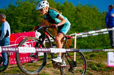 Junge Radfahrerin in Helm und Sportkleidung fährt Mountainbike auf Rennstrecke mit Absperrband und Zuschauern im Hintergrund
