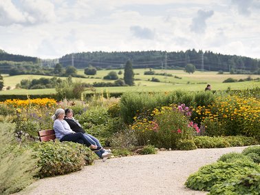 Two people sitting on a bench in a garden with colorful flowers and rolling hills in the background