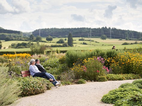 Zwei ältere Frauen sitzen auf einer Bank in einem blühenden Garten mit Blick auf grüne Felder und bewaldete Hügel im Hintergrund
