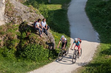 Zwei Mountainbiker fahren einen Schotterweg entlang, während weitere zwei Personen auf einem Felsen sitzen.