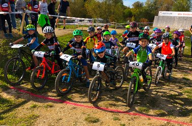 Kinder starten ein Mountainbike-Rennen auf einem Waldweg, alle tragen Helme und bunte Sportkleidung