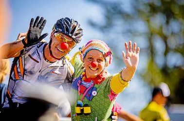 Zwei Radfahrer mit Helmen und roten Clownnasen winken in die Kamera bei sonnigem Wetter