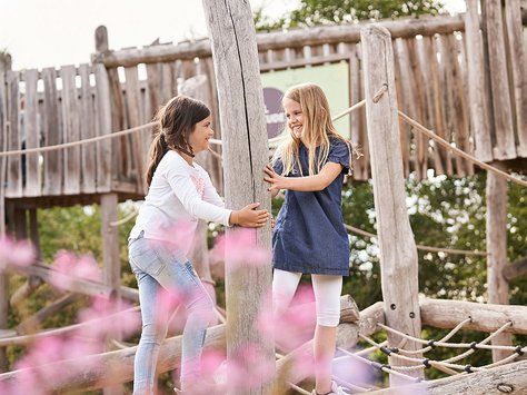 Zwei Mädchen spielen lachend auf einem Holzspielplatz im Freien, umgeben von grünen Bäumen und rosa Blumen