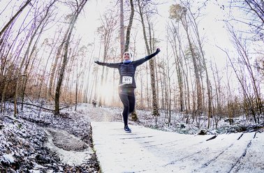 Läufer mit ausgestreckten Armen auf verschneitem Waldweg, umgeben von kahlen Bäumen
