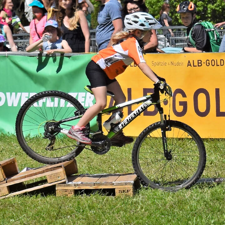 Mountainbiker in orangefarbenem Trikot fährt über eine Holzkonstruktion bei einem Wettkampf im Freien