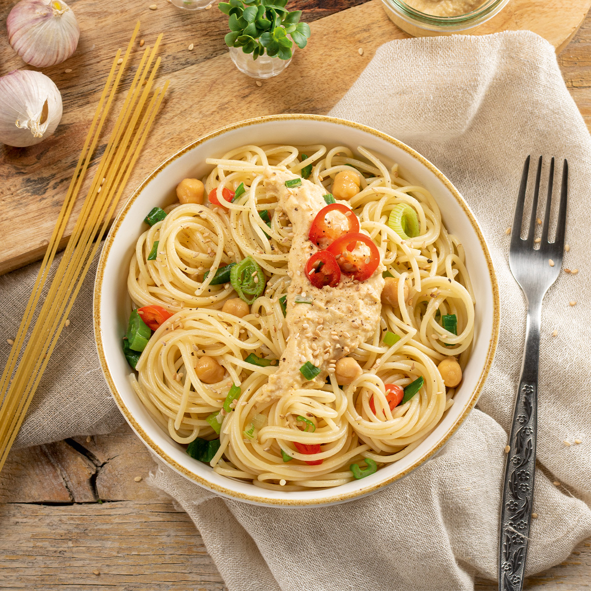 Schüssel mit Spaghetti, Kichererbsen, Kirschtomaten und Kräutern auf einem Leinentuch, daneben Gabel und rohe Spaghetti