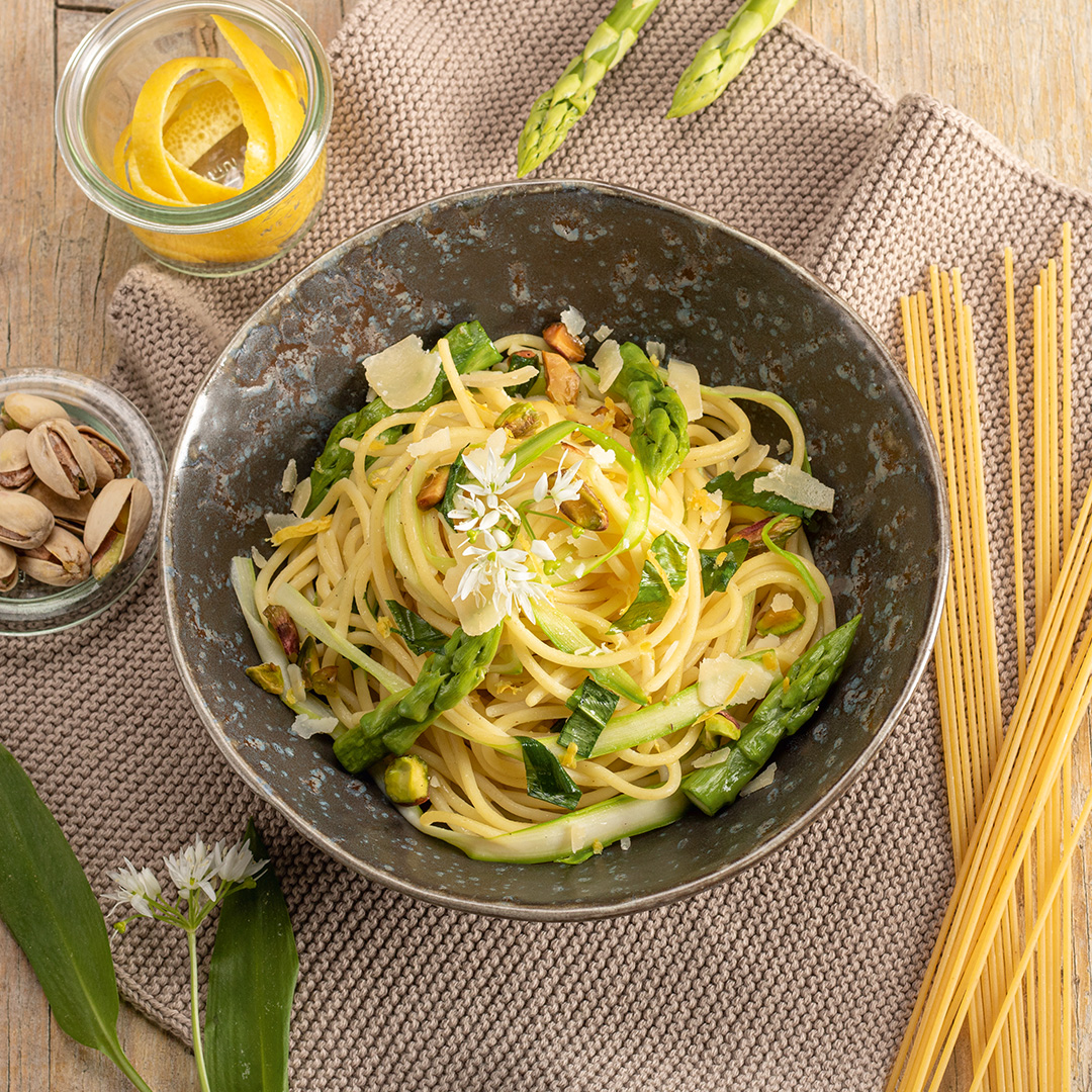 Schüssel mit Spaghetti, Spargelstücken und Parmesan auf einem Tisch mit rohem Spargel und ungekochten Spaghetti
